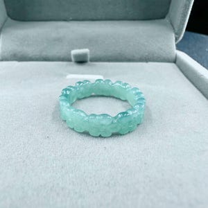 Puede incluir: Un anillo verde claro con un diseño floral. El anillo está hecho de un material translúcido y se muestra en una caja de joyería gris. El diseño del anillo presenta una serie de formas de flores conectadas.