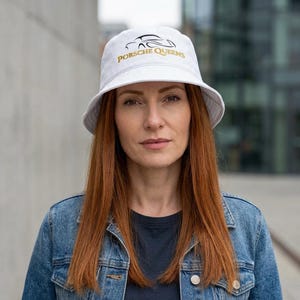 May include: White bucket hat with the text "PORSCHE QUEENS" in gold and a black car outline. The hat is made of a soft material and has a wide brim. The hat is worn by a person with long red hair and a denim jacket.