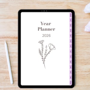 May include: A digital year planner for 2026 displayed on a black tablet. The planner's cover features the text "Year Planner 2026" and a floral illustration. A white stylus rests on the wooden surface beside the tablet. The planner has monthly tabs.