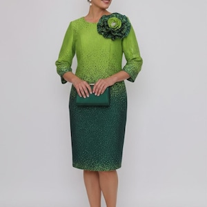 以下が含まれることがあります： テクスチャード加工のパターンが施されたグリーンのグラデーションドレス。肩には大きな花のアクセントが付いています。ドレスは七分袖で、お揃いのグリーンのクラッチとヒールが組み合わされています。全体的にエレガントで洗練された印象です。