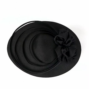 Peut inclure: Un chapeau noir avec un large bord et un accent floral d&eacute;coratif. Le chapeau pr&eacute;sente un design circulaire avec des bandes superpos&eacute;es et une fleur en tissu sur le c&ocirc;t&eacute;. Le chapeau est fait d'un mat&eacute;riau textur&eacute;.
