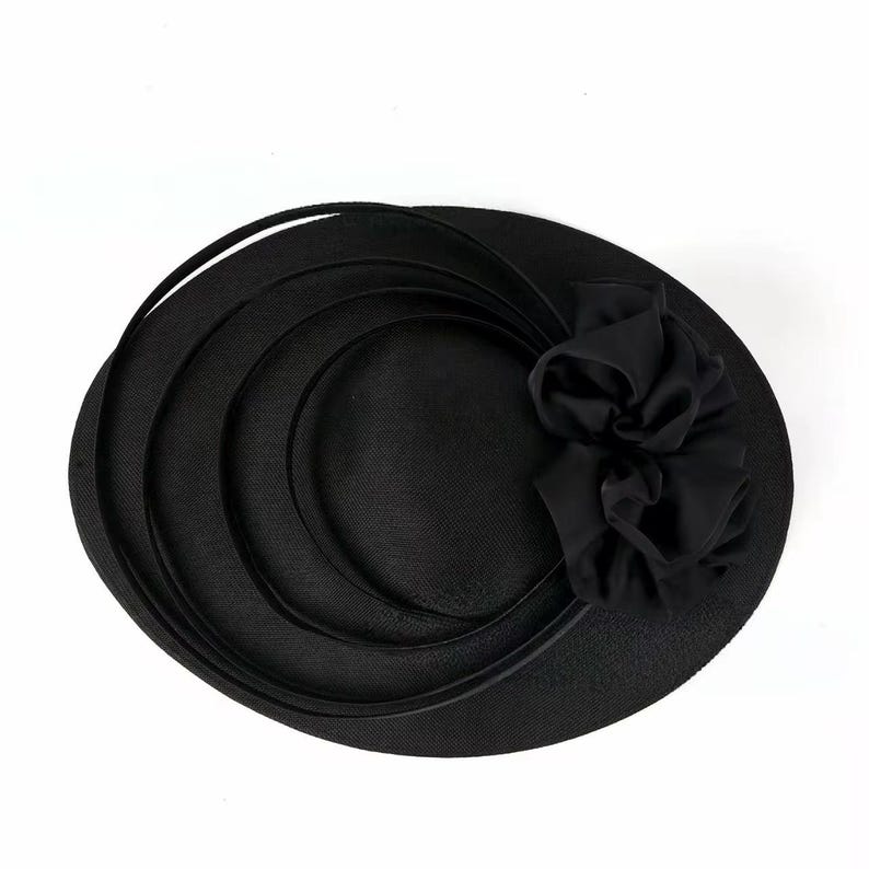 Peut inclure: Un chapeau noir avec un large bord et un accent floral d&eacute;coratif. Le chapeau pr&eacute;sente un design circulaire avec des bandes superpos&eacute;es et une fleur en tissu sur le c&ocirc;t&eacute;. Le chapeau est fait d'un mat&eacute;riau textur&eacute;.