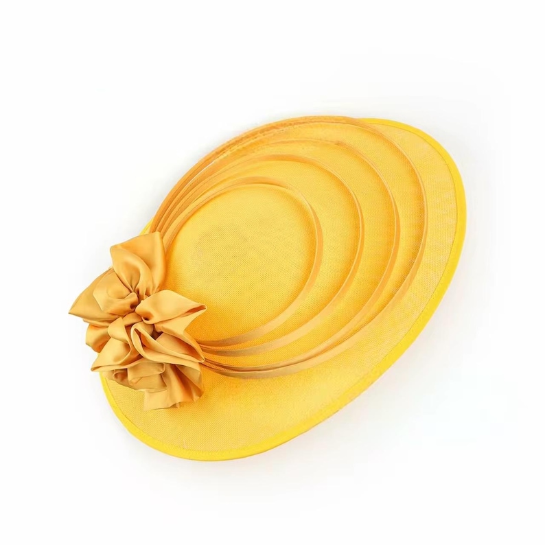 Peut inclure: Un chapeau jaune vif de forme ovale, orn&eacute; d'un n&oelig;ud d&eacute;coratif sur le c&ocirc;t&eacute;. Le chapeau pr&eacute;sente des bandes incurv&eacute;es concentriques et une texture en forme de filet. Le n&oelig;ud est en tissu jaune assorti.