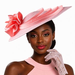 May include: A light pink hat with a large, ruffled flower on the side. The hat has a wide brim and is adorned with ribbons. The woman in the image is wearing a matching pink dress and lace gloves, creating a coordinated look.