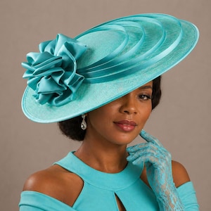 Peut inclure: Un chapeau turquoise &agrave; large bord orn&eacute; d'une grande fleur froiss&eacute;e sur le c&ocirc;t&eacute;. Le chapeau pr&eacute;sente des rubans superpos&eacute;s et est fait d'un tissu textur&eacute;. La femme porte une robe et des gants en dentelle turquoise assortis.