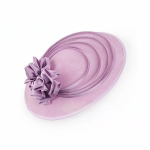 Peut inclure: Un chapeau de c&eacute;r&eacute;monie de couleur lavande avec une grande fleur froiss&eacute;e sur le c&ocirc;t&eacute;. Le chapeau pr&eacute;sente un design circulaire avec des accents incurv&eacute;s superpos&eacute;s. Le bord est bord&eacute; d'une garniture lavande assortie. Coiffe &eacute;l&eacute;gante pour occasions sp&eacute;ciales.