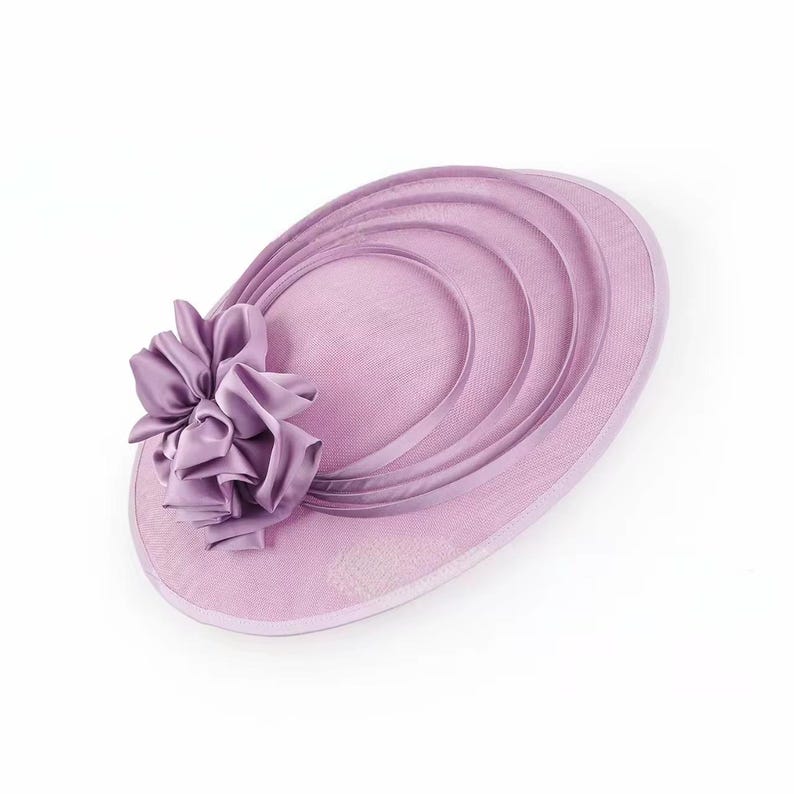 Peut inclure: Un chapeau de c&eacute;r&eacute;monie de couleur lavande avec une grande fleur froiss&eacute;e sur le c&ocirc;t&eacute;. Le chapeau pr&eacute;sente un design circulaire avec des accents incurv&eacute;s superpos&eacute;s. Le bord est bord&eacute; d'une garniture lavande assortie. Coiffe &eacute;l&eacute;gante pour occasions sp&eacute;ciales.
