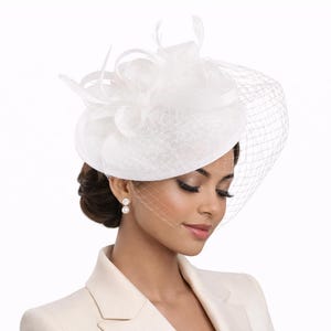 Op de afbeelding: Een witte formele hoed met sluier en verenaccenten. De hoed heeft een ronde kroon en een fijn gaas dat over het gezicht valt. De hoed is gemaakt van een textuurstof en is geschikt voor speciale gelegenheden.