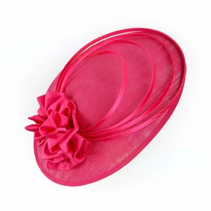 Peut inclure: Un chapeau de c&eacute;r&eacute;monie fuchsia vif avec une grande fleur froiss&eacute;e sur le c&ocirc;t&eacute;. Le chapeau pr&eacute;sente une base en r&eacute;sille et des rubans d&eacute;coratifs, id&eacute;al pour les occasions sp&eacute;ciales. Forme ovale.