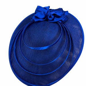 Peut inclure: Un chapeau bleu roi avec un grand n&oelig;ud et trois cercles concentriques. Le chapeau a un large bord et un tissage textur&eacute;. Le n&oelig;ud est fait d'un tissu lisse et brillant. Un accessoire de c&eacute;r&eacute;monie.