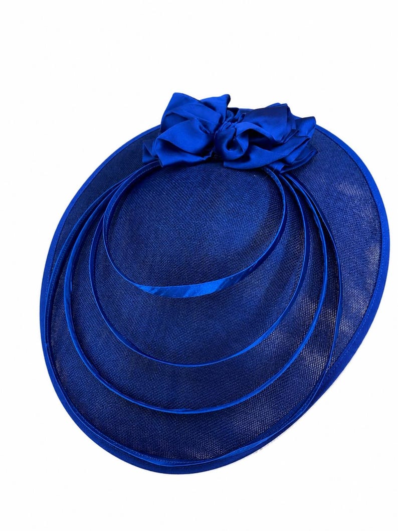 Peut inclure: Un chapeau bleu roi avec un grand n&oelig;ud et trois cercles concentriques. Le chapeau a un large bord et un tissage textur&eacute;. Le n&oelig;ud est fait d'un tissu lisse et brillant. Un accessoire de c&eacute;r&eacute;monie.