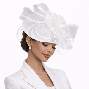 May include: A white fascinator hat featuring a structured base, large layered organza loops, and a central floral accent. The design includes a delicate netting veil that extends over the side, creating a formal headpiece with a clean, monochromatic aesthetic.