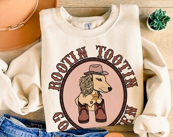 May include: Cream-colored sweatshirt with a circular graphic featuring a cartoon dog wearing a cowboy hat and boots. The text "Rootin' Tootin' Go Hen" surrounds the dog illustration. A brown hat and blue jeans are visible.