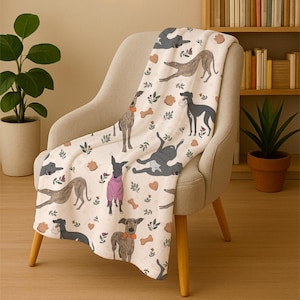 May include: A cream-colored throw blanket draped over a chair, featuring a repeating pattern of illustrated greyhounds in various poses. The design includes dog bones, hearts, and floral accents. The blanket is made of a soft material.