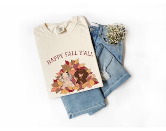 May include: Cream-colored t-shirt with the text "HAPPY FALL Y'ALL" above a graphic of two dachshunds in a pile of autumn leaves. The shirt is paired with blue denim shorts and a small bouquet of white flowers.
