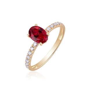 May include: A gold ring featuring a large, oval-cut, deep red gemstone as the centerpiece. The band is adorned with small, clear gemstones. The ring is set against a white background, highlighting its elegant design.