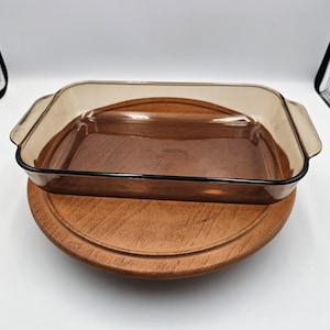 May include: A rectangular amber-tinted glass baking dish with integrated side handles. The dish features a transparent brown hue and is positioned on a circular wooden base. The bottom of the glass dish has embossed text that is partially visible.