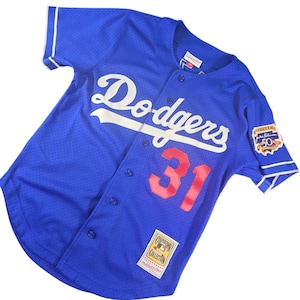Può includere: Una maglia da baseball blu dei Dodgers con la scritta "Dodgers" in bianco sul petto e il numero "31" in rosso. La maglia ha un'abbottonatura frontale e una toppa "Jackie Robinson 50" sulla manica.