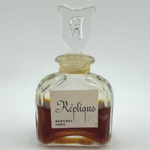 May include: A clear glass perfume bottle with a decorative stopper. The bottle has a square shape with rounded edges and contains a dark amber liquid. The label reads "Réplique RAPHAEL PARIS". The stopper is clear glass with the letter "R".