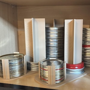 May include: A collection of silver metal jar lids and red-banded lids, organized with white dividers. The lids are stacked in neat piles, with some held together by wooden supports. The text "Wide Mouth" is visible on the dividers.