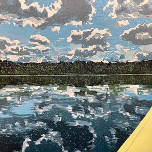 May include: A landscape print depicts a serene lake scene. The sky is filled with fluffy white and gray clouds against a light blue backdrop. The water reflects the sky and the dark green treeline in the distance. A yellow kayak is in the foreground.