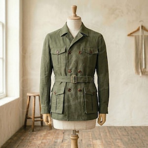 Puede incluir: Chaqueta verde oliva con diseño de doble botonadura, cuatro bolsillos delanteros y cinturón a juego. La chaqueta tiene botones marrones y se exhibe en un maniquí. El estilo recuerda a una prenda de inspiración safari o militar.