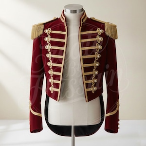 Op de afbeelding: Bordeaux jas in militaire stijl met gouden biezen, epauletten en parelachtige knoopaccenten. De jas heeft een hoge kraag en een jacquet-ontwerp. De gouden biezen omlijnen de randen, manchetten en decoratieve elementen, wat een opvallend contrast creëert.