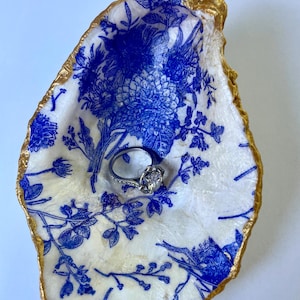 Puede incluir: Un plato para anillos con forma de concha de ostra decorado con un estampado floral azul sobre fondo blanco. El borde está pintado de dorado. Un anillo de plata con una piedra ovalada descansa dentro del plato.
