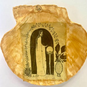 May include: A decorative seashell with a gold-colored exterior. The shell holds a rectangular image featuring a black and white illustration of a woman in a long gown, a crown, and a sphere. The image is framed by an arch and botanical elements.