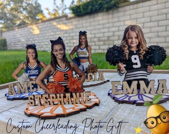 May include: Four custom photo cutouts of cheerleaders mounted on wooden bases. Each base features a name in wooden letters: Amelia, Stephanie, Alicia, and Emma. The cheerleaders are wearing uniforms and holding pom-poms in various colors.