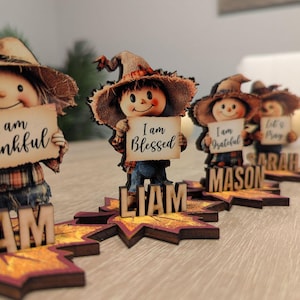 May include: A collection of wooden scarecrow figurines, each holding a sign with a positive message. The figurines have straw hats and are placed on leaf-shaped bases with names. The signs read "I am Thankful", "I am Blessed", "I am Grateful", and "Let's Pray".