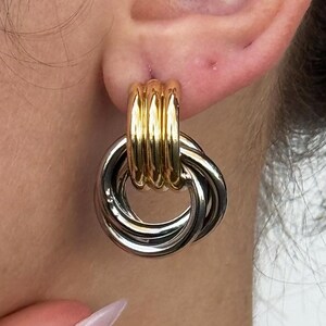 May include: Gold and silver-toned hoop earrings. The earrings feature a gold-colored post with a series of vertical lines, and a silver-colored interlocking hoop design. The earrings are a statement piece.