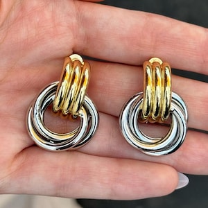 May include: A pair of gold and silver-toned earrings. Each earring features a gold-colored, stacked, cylindrical top and a silver-colored, interlocking circular design. The earrings are displayed against a neutral background.