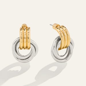 May include: A pair of two-tone earrings. The top portion is gold-colored, featuring three parallel bars. The lower section is silver-colored, with two interlocking oval rings. The earrings are displayed on a light background.
