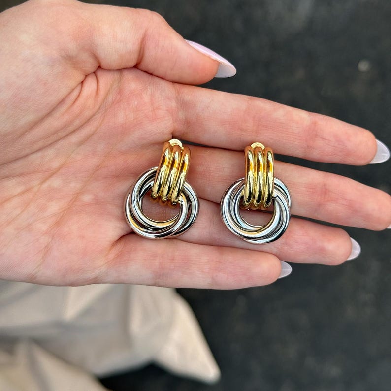 May include: A pair of gold and silver-toned earrings. Each earring features a gold-colored, stacked, cylindrical top and a silver-colored, interlocking circular design. The earrings are displayed against a neutral background.