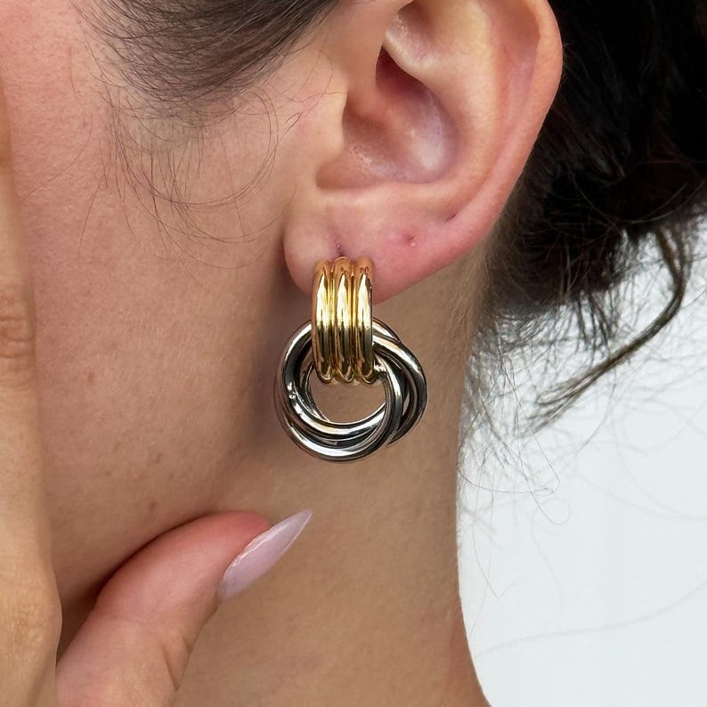 May include: Gold and silver-toned hoop earrings. The earrings feature a gold-colored post with a series of vertical lines, and a silver-colored interlocking hoop design. The earrings are a statement piece.
