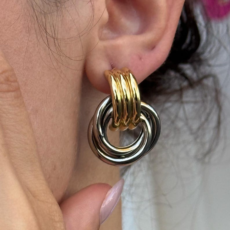 May include: Gold and silver-tone earrings with a linked ring design. The earrings feature a gold-colored top section with three vertical bars, and three interlocking silver-colored rings below. The earrings are shown on an ear.