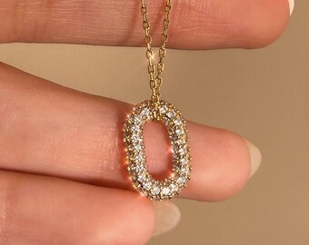 May include: A gold-colored necklace with a pendant shaped like an oval. The oval is encrusted with small, clear gemstones. The chain is delicate and also gold-colored. The necklace is held up by a person's fingers.