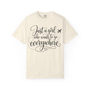 May include: A cream-coloured short-sleeve t-shirt featuring black cursive text that reads Just a girl who wants to go everywhere. The design includes a small aeroplane icon with a dashed flight path line and a heart symbol integrated into the lettering.
