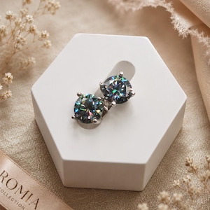 May include: A pair of round-cut stud earrings featuring dark, iridescent stones with multi-colored light reflections, set in a silver-toned metal prong setting. The earrings are displayed on a white hexagonal stand with the text ROMIA COLLECTION.