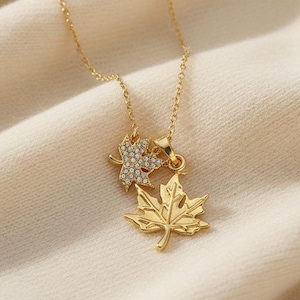 Peut inclure: Un collier doré avec deux pendentifs en forme de feuille d'érable. L'un est une feuille pleine et détaillée, l'autre une petite feuille sertie de cristaux transparents. La chaîne fine est posée sur un tissu crème.