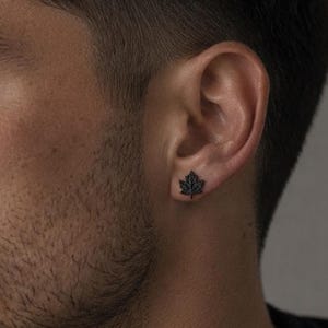 以下が含まれることがあります： 詳細なカエデの葉の形をした小さなマットブラックのスタッドピアス。ピアスは耳たぶに装着されており、自然の葉の葉脈や尖った端を模した質感のある表面が特徴で、控えめでダークな美学を提供しています。