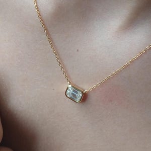 Puede incluir: Un collar de oro con un colgante rectangular de piedra preciosa transparente. El colgante está engastado en un bisel de oro y cuelga de una delicada cadena de oro. El collar se muestra sobre un fondo de tono de piel.
