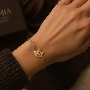 May include: A gold-toned chain bracelet featuring a central maple leaf charm with cut-out detailing. The delicate metal chain connects to the leaf on both sides, creating a simple and refined piece of jewellery worn around the wrist.