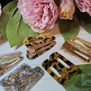 Könnte beinhalten: Eine Auswahl an Haarspangen in verschiedenen Designs und Farben. Die Spangen weisen Schildpatt-, Gold- und transparente Muster auf. Einige sind rechteckig, andere oval. Rosa Blumen und grüne Blätter im Hintergrund.