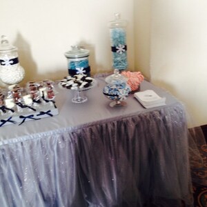May include: A table covered in a silver sparkly tablecloth with a variety of candy jars and treats. The jars are filled with blue and white candies and the treats are chocolate covered pretzels on sticks.