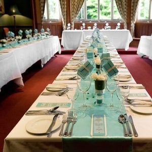 May include: A long table set for a wedding reception with white tablecloths and teal table runners. The centerpieces are small boxes tied with teal ribbon and white flowers. The table is set with white plates, silverware, and glasses.