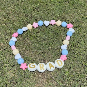 Puede incluir: Una guirnalda de bolas y estrellas de color pastel azul, rosa y crema con la palabra "GRAD" escrita en letras blancas con purpurina dorada.