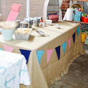 May include: A burlap table cloth with a ruffled edge is decorated with a blue, pink, and white pennant banner. The table is set with a variety of food and drinks, including a bucket of wooden skewers, a bowl of french fries, and a variety of plates and bowls.