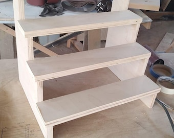 May include: A four-tiered wooden display stand made from light-coloured plywood. The stand features a stepped design with four shelves of varying heights. It is designed for displaying items and has a simple, functional construction.